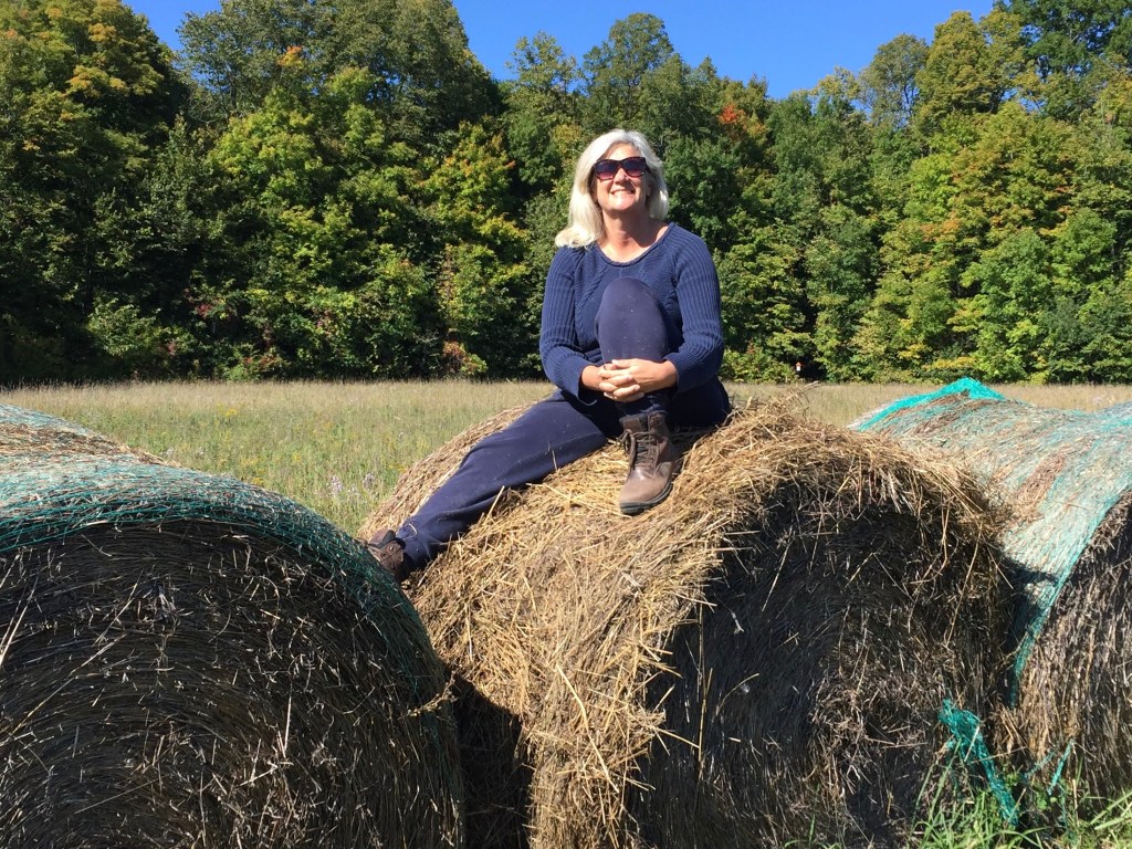 Arlene sitting on a round hay bale in a farm field. 