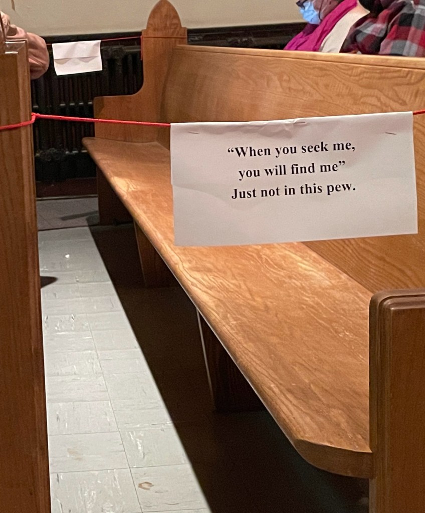 An empty row of seating in a church. A sign hangs from a barrier blocking access to the row. The sign reads, "'When you seek me, you will find me.' Just not in this pew."