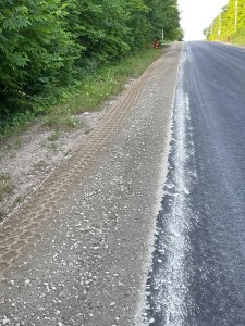View up the hill of a country road with new gravel shoulders and fresh pavement. Construction pylons frame the road at the top of the hill.
